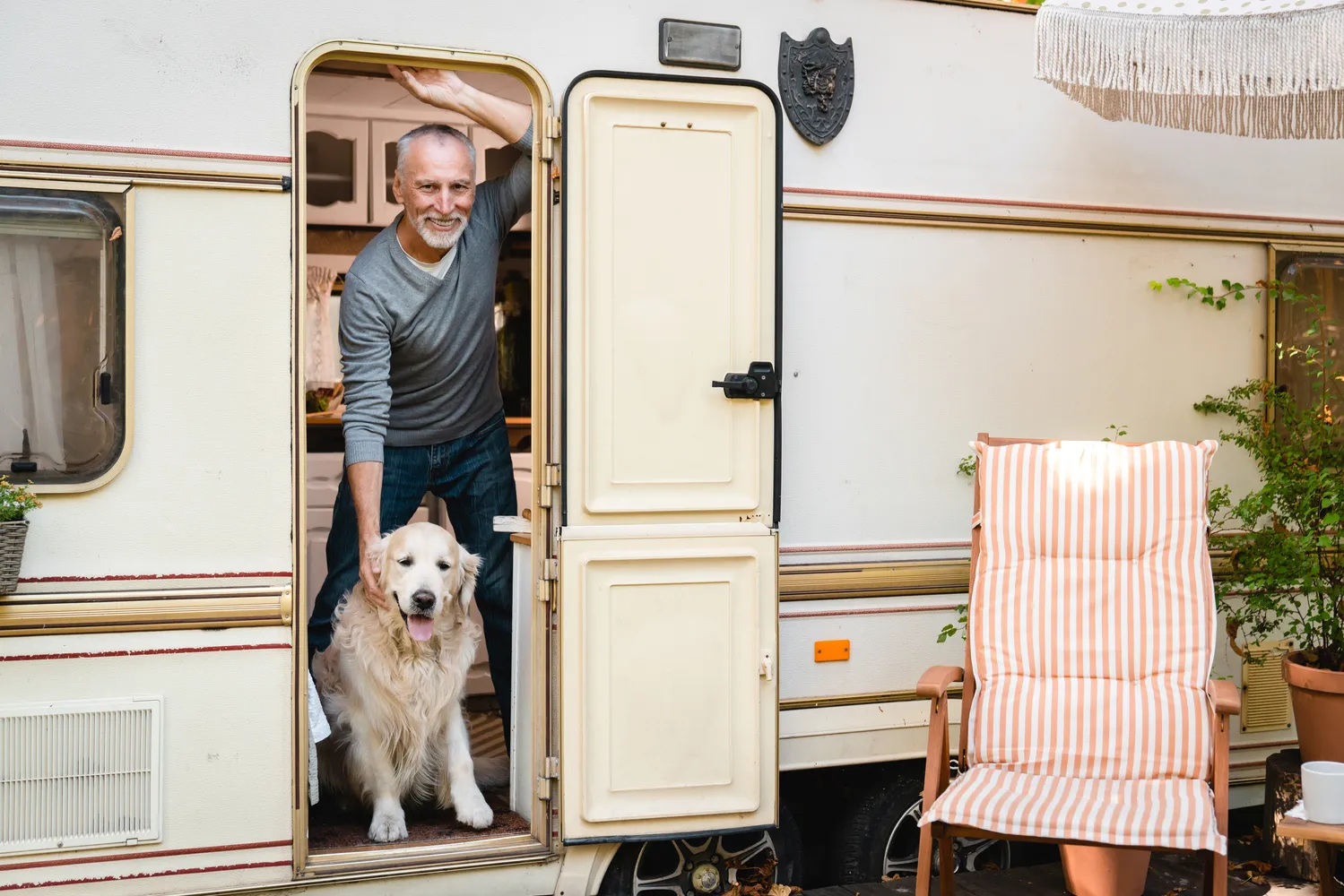 Happy older gentleman leaning in the doorway of a classic cream-colored travel trailer with his friendly golden labrador – get personalized coverage for your camper and lifestyle from a reliable insurance agency.