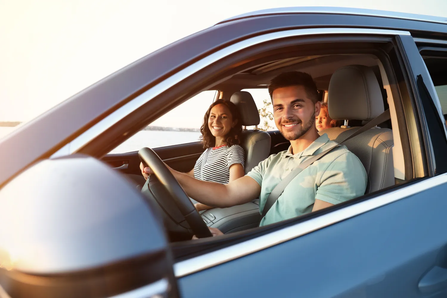 Joyful parents and young daughter buckled up in a blue crossover car, laughing during a peaceful evening drive by the water – secure your loved ones on the road with personalized auto insurance from an experienced insurance agent.