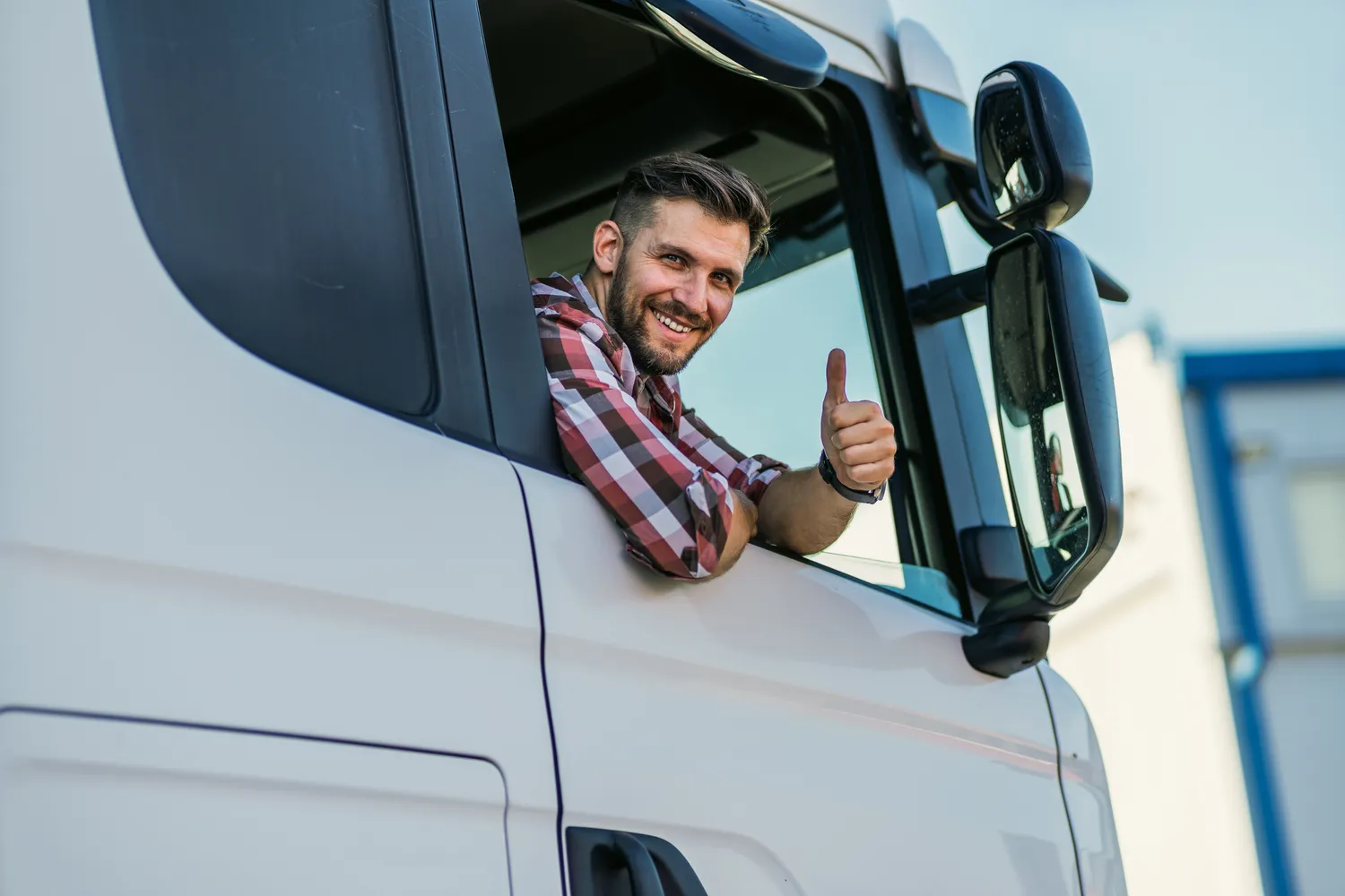 Smiling truck driver leaning out of cab window giving thumbs up in front of white semi-truck – protect your commercial fleet and livelihood with specialized truck insurance from a trusted insurance agent.
