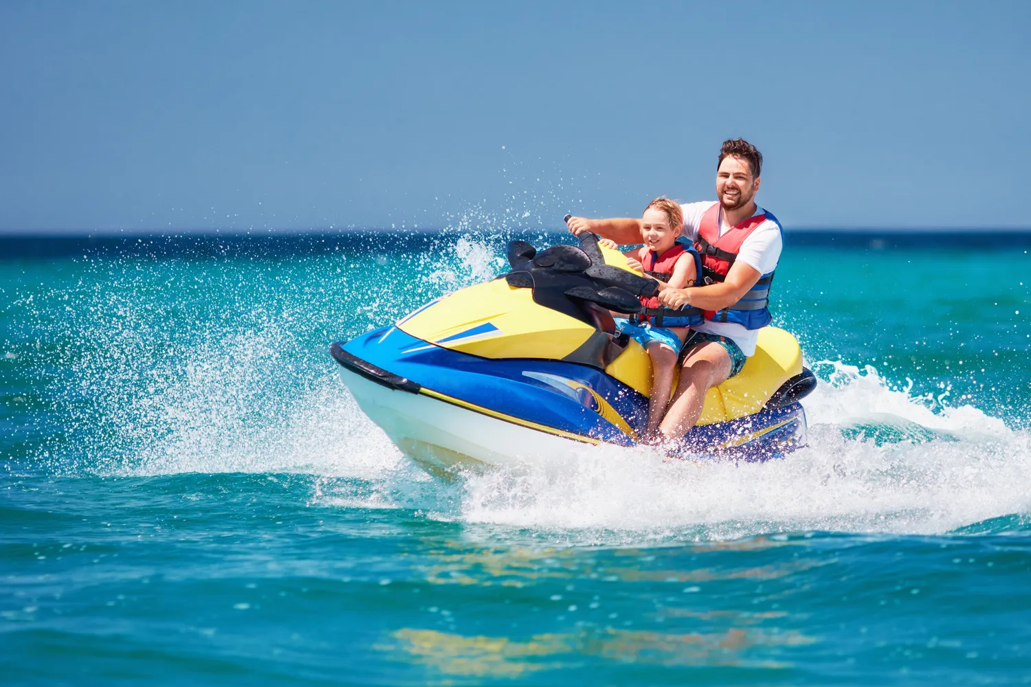 Father and young son riding a vibrant yellow and blue jet ski together on turquoise ocean waves – protect your family water sports with expert advice from a trusted insurance agent.